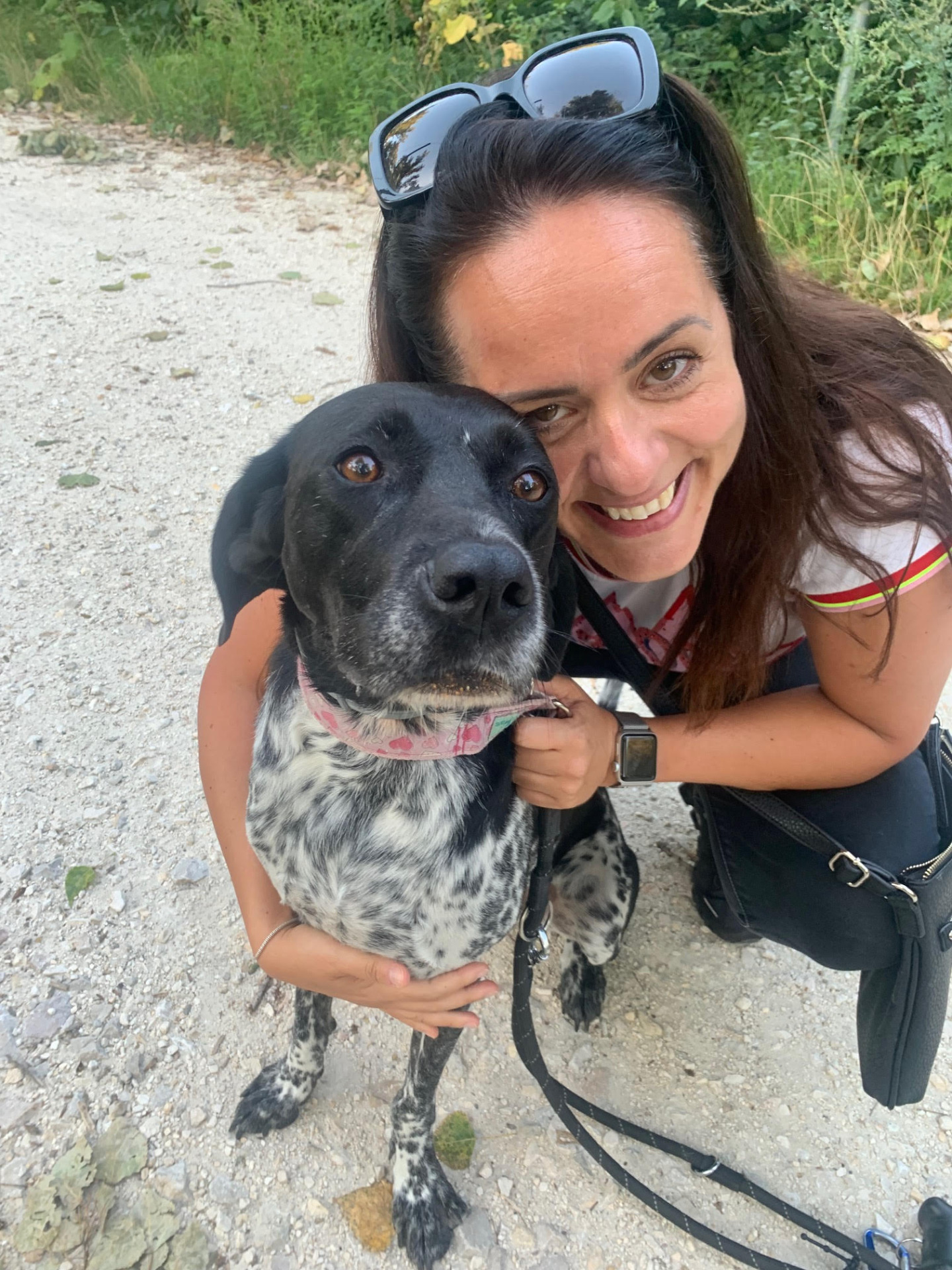 Smiling woman holds a black-and-white dog outdoors on a gravel path with greenery behind.