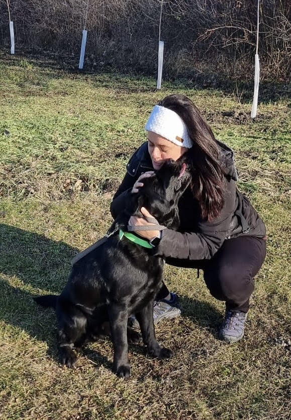 Marta, the dog trainer, kneels in a grassy field, hugging a black labrador dog wearing a green collar.